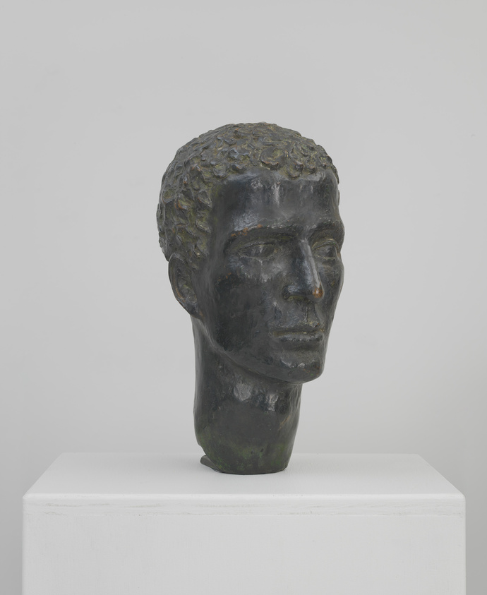 A bronze sculpture of a human head with textured hair, displayed on a white pedestal against a light background.