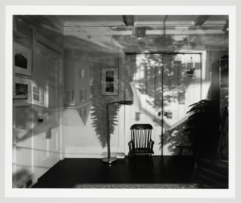 Black and white photograph of an interior room with a rocking chair, framed pictures on the wall, and a floor lamp. The room is overlaid with shadows of trees and the exterior of a building, suggesting a double exposure or reflection effect.