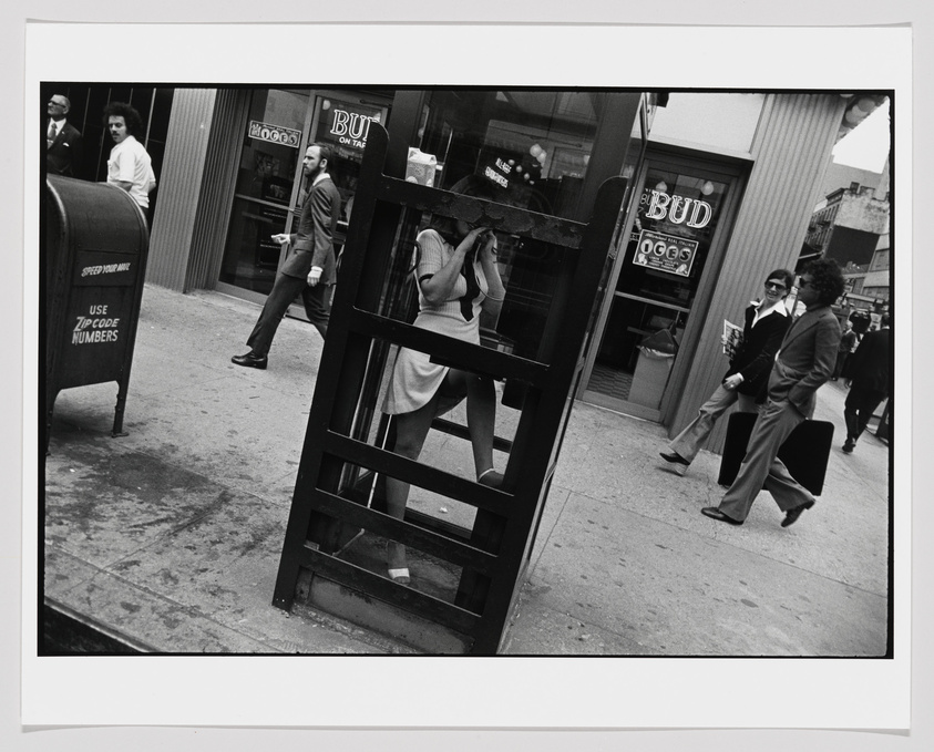 A black and white street photograph capturing a moment in time with various people walking by. In the foreground, a woman is seen through a glass door, her face obscured by her hand and the door frame. A man in a suit walks past a mailbox that reads "SPEED YOUR MAIL USE ZIP CODE NUMBERS." In the background, other individuals are seen walking, including a couple in conversation. The scene includes a bar with a "BUD" beer sign in the window, suggesting an urban setting. The photo is framed with a white border.