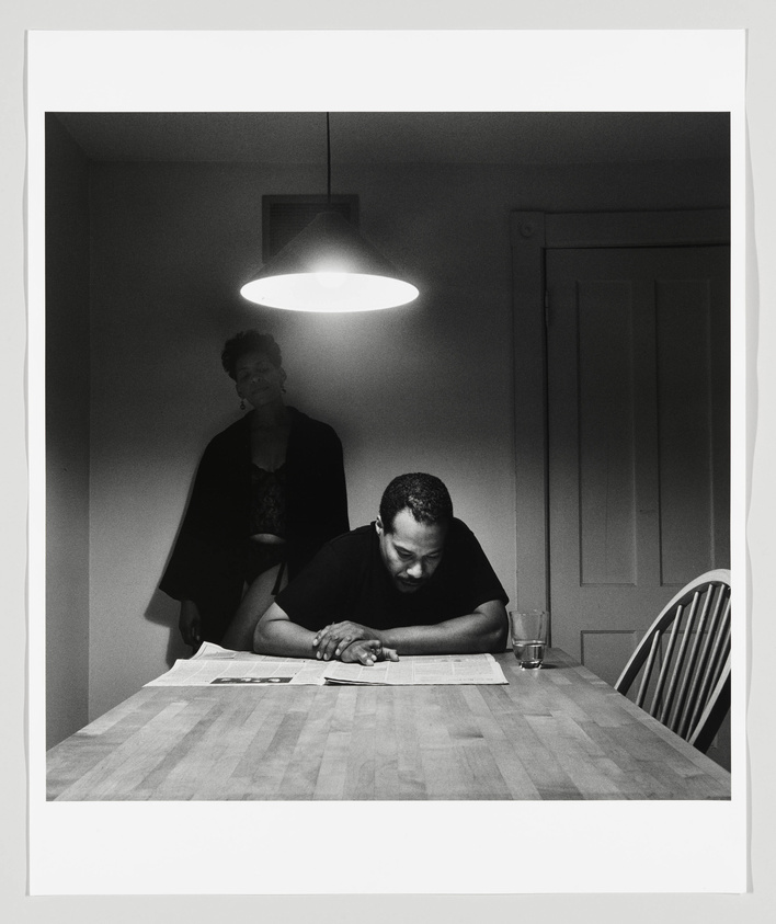 A black and white photograph depicting a man sitting at a wooden table, reading a newspaper under a hanging lamp, with a woman standing in the background observing him. The room appears to be a kitchen or dining area with a closed door on the right. The lighting creates a dramatic atmosphere, highlighting the subjects and the table.