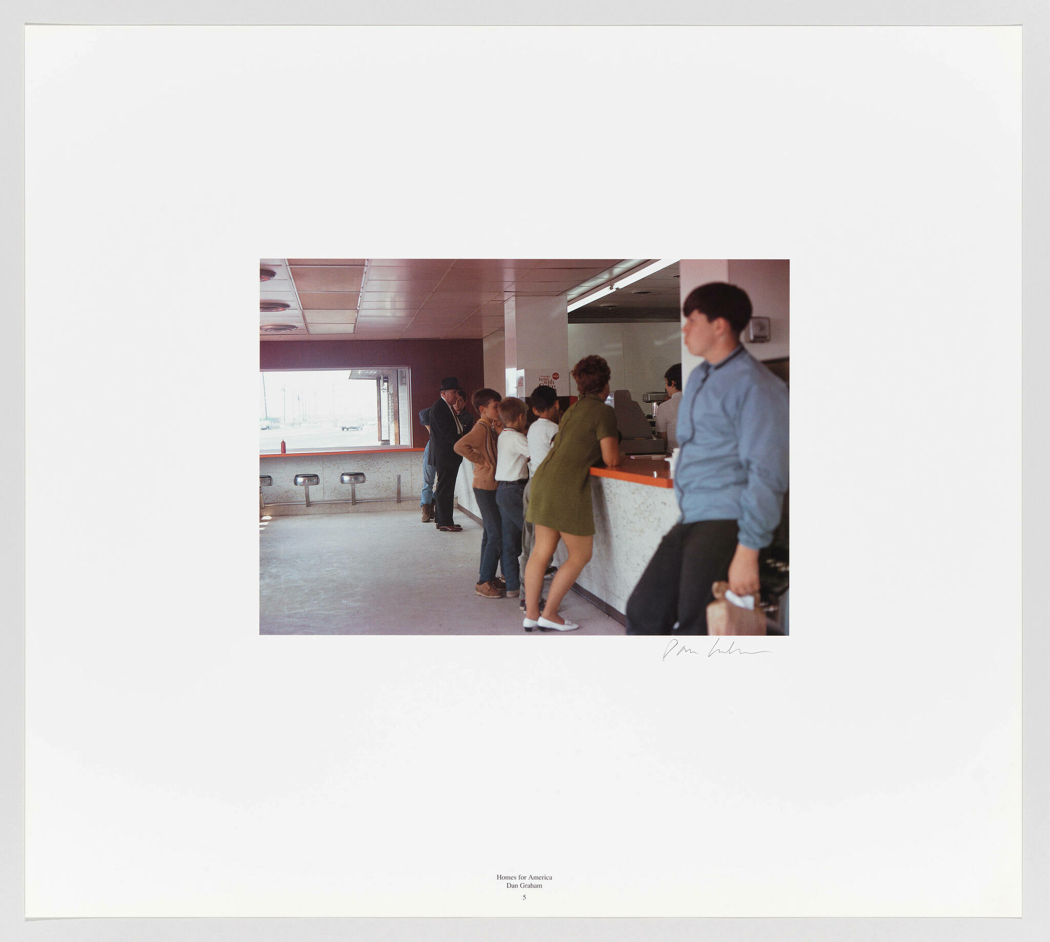 A photograph of a group of people standing in line at a counter, with a man in the foreground looking to the side. The setting appears to be an indoor public space with a minimalist design and large windows in the background. The image is framed with a wide white border that includes the title "Homes for America" and the name Dan Graham at the bottom, along with a signature on the right.