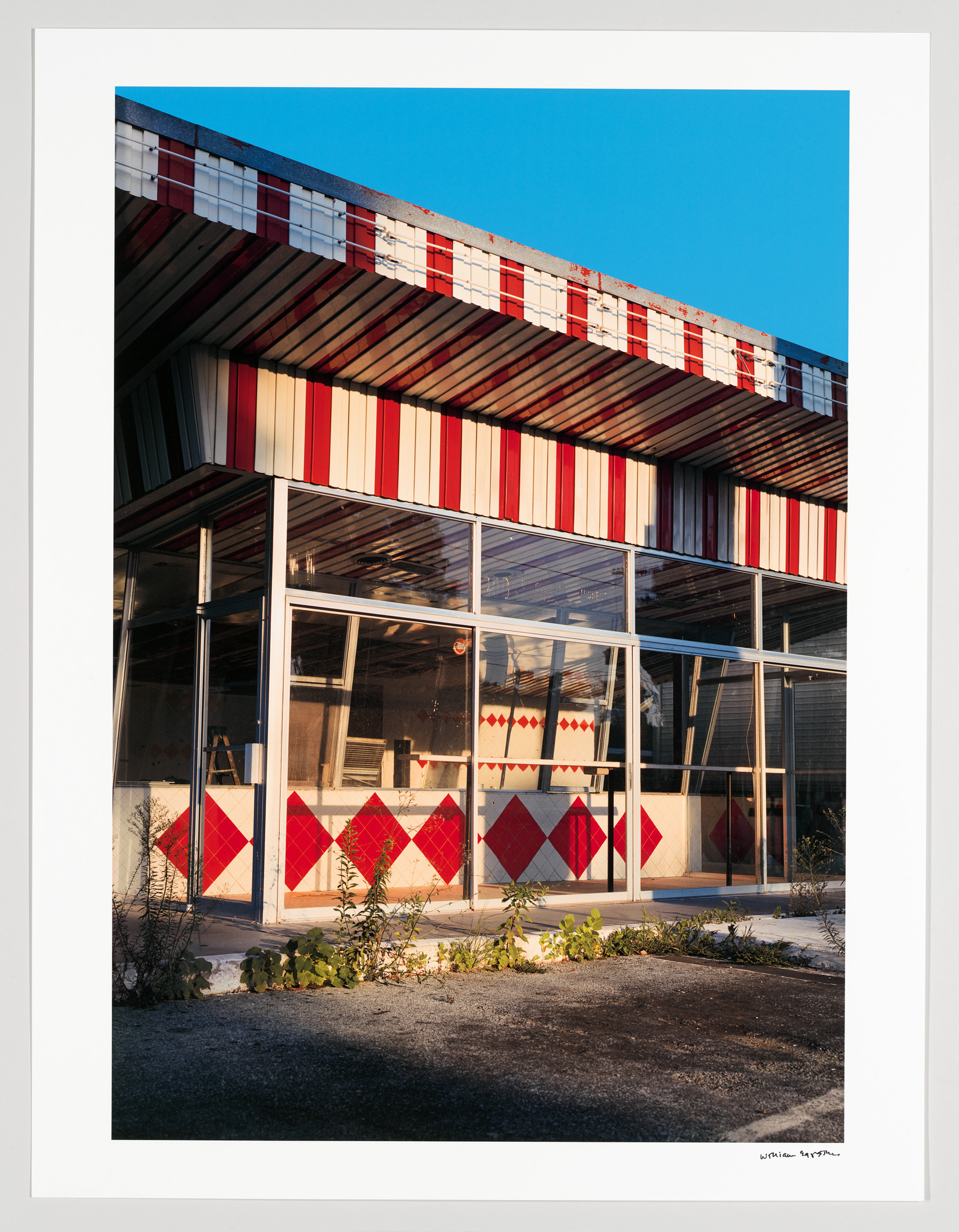 A modern building with a geometric design featuring a pattern of red and white stripes and diamond shapes. The structure has large glass windows and is illuminated by warm sunlight, with a clear blue sky in the background. The image is framed with a white border and includes a signature in the bottom right corner.