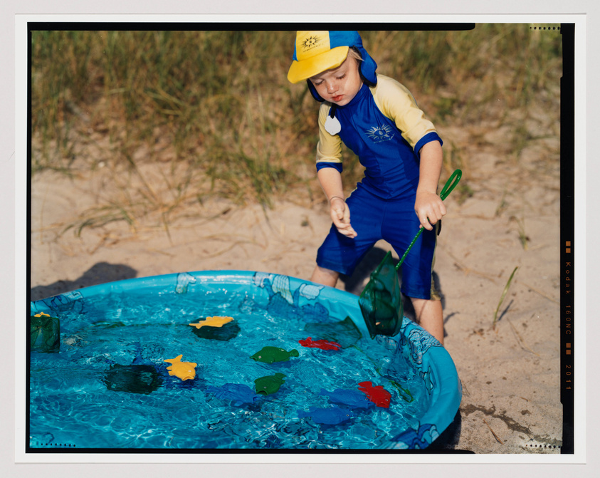 A young child wearing a blue and yellow swimsuit with a matching hat is playing with a green net in a small inflatable pool filled with water and floating plastic toys. The pool is set on a sandy area with grass in the background, suggesting a beach setting. The image has a film border with exposure and color balance markings, indicating it may be a photograph or styled to look like one.
