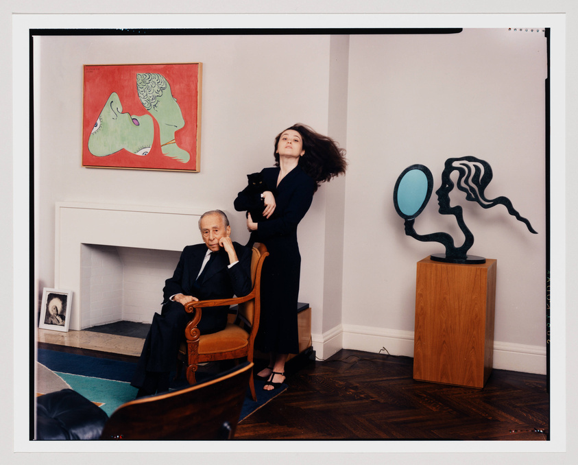 A photograph of an older man seated in a wooden armchair, looking contemplatively to the side, with a younger woman standing behind him, holding a black cat and looking off into the distance. They are in a room with modern art on the walls, including a red painting with two green profiles and a sculpture of a stylized figure with a mirror for a face. A fireplace and a framed photo on the mantel are also visible.