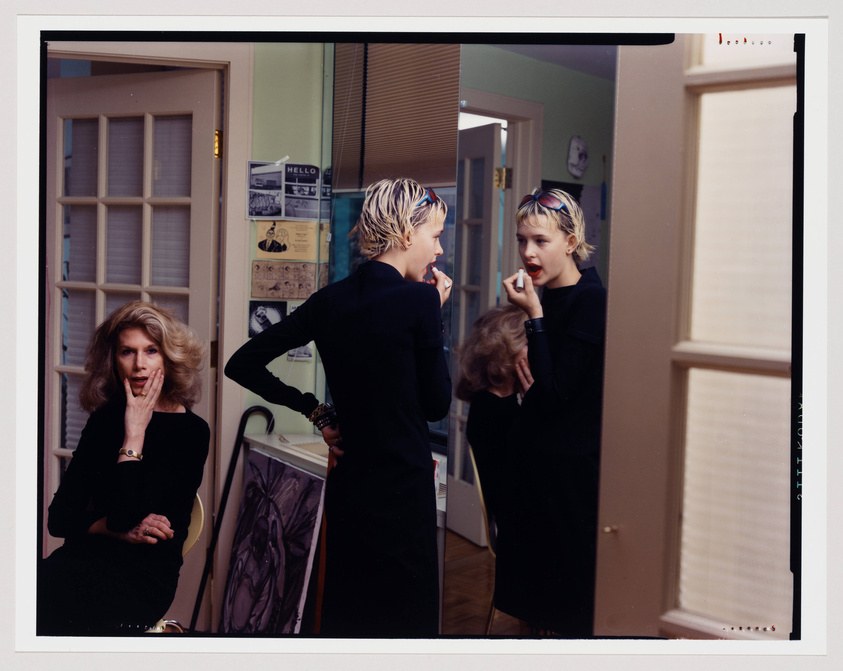 A staged photograph capturing three women in a domestic setting. On the left, a woman with shoulder-length blonde hair, wearing a black dress and a watch, sits with her hand to her cheek, looking surprised or contemplative. In the center, a young woman with short blonde hair, dressed in black with a colorful headband, is applying red lipstick in front of a mirror, her reflection visible. On the right, the reflection shows the young woman's back and the side of her face as she focuses on her makeup. The room has a retro feel with a green wall, a bulletin board with various notes, and a glimpse of a window with blinds.