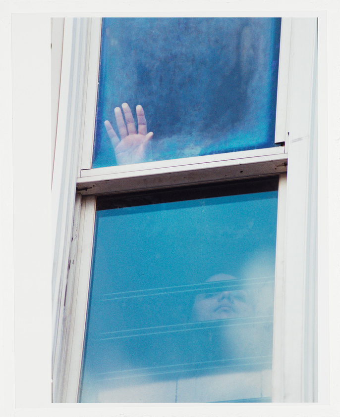 A photograph showing a double window with the upper pane frosted. A person's hand is pressed against the inside of the frosted glass, while the lower clear pane shows a reflection of another person's face looking upward. The window frame is white, and the image has a slightly bluish tint.