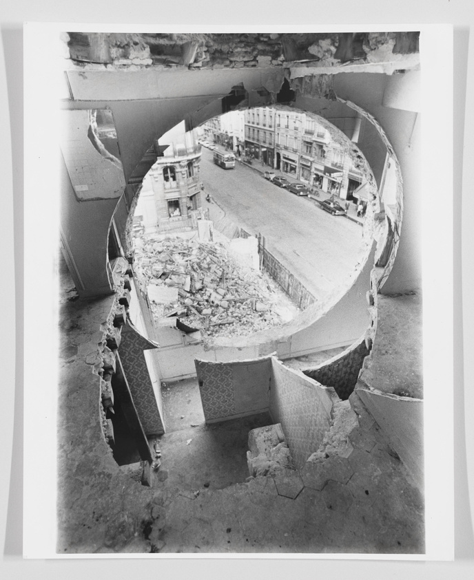 A black and white photograph showing a view through a large, circular hole in a damaged building wall, overlooking a street scene with buildings, cars, and a bus. Debris and broken walls are visible in the foreground, indicating a scene of destruction or demolition.