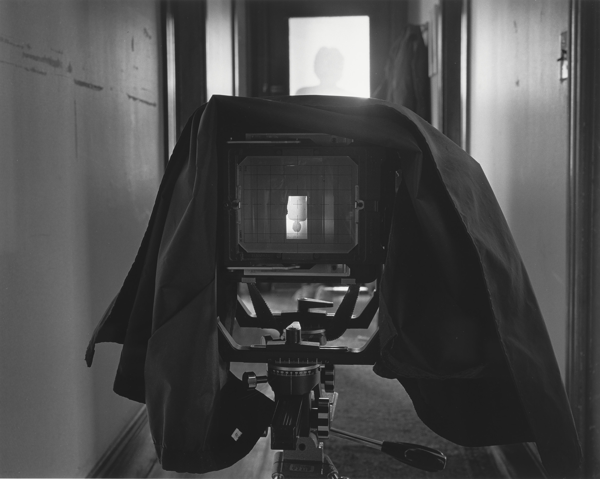Large format camera on tripod in hallway, focused on a silhouetted figure in the distance.