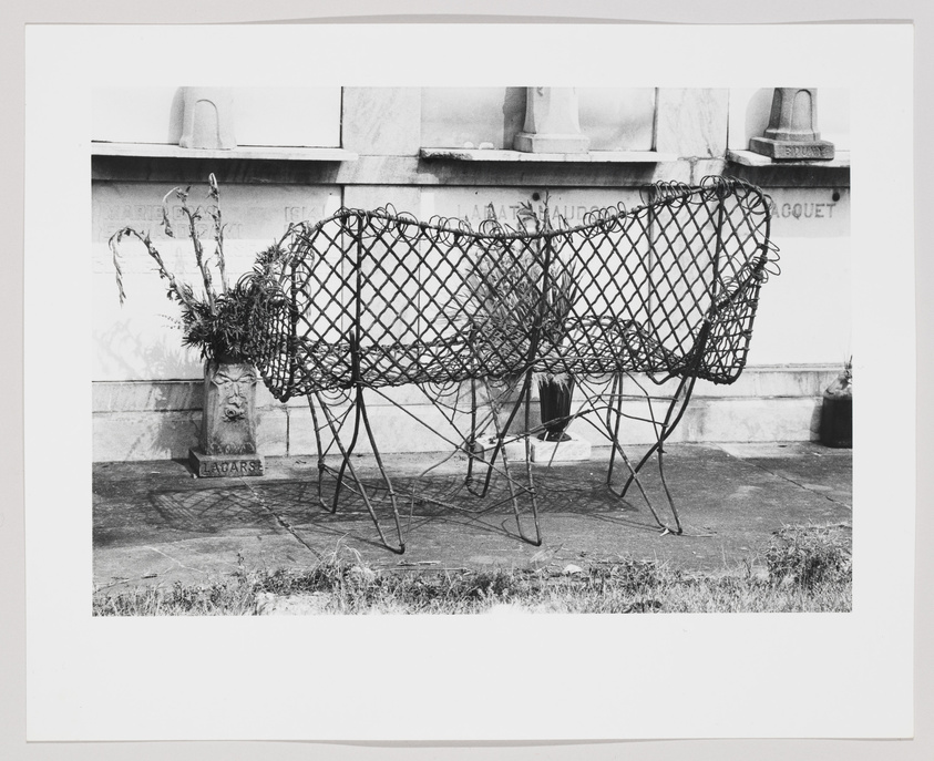 A black and white photograph depicting a wireframe sculpture of a sheep with a flower arrangement on its back, standing in front of a wall with inscriptions and other grave markers in a cemetery.