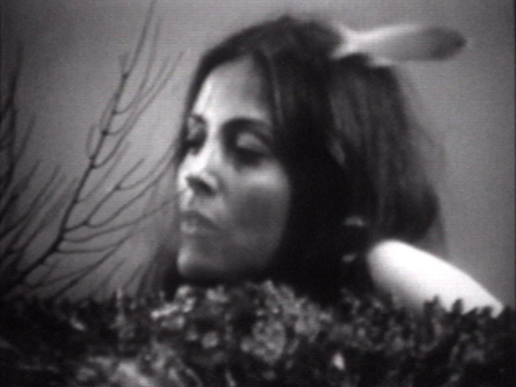 Black and white image of a woman lying down with her eyes closed, appearing peaceful or asleep. Her head is resting on a pillow, and there is a blurred foreground that might be a floral arrangement. The image has a grainy texture, suggesting it might be from an older film or television broadcast.