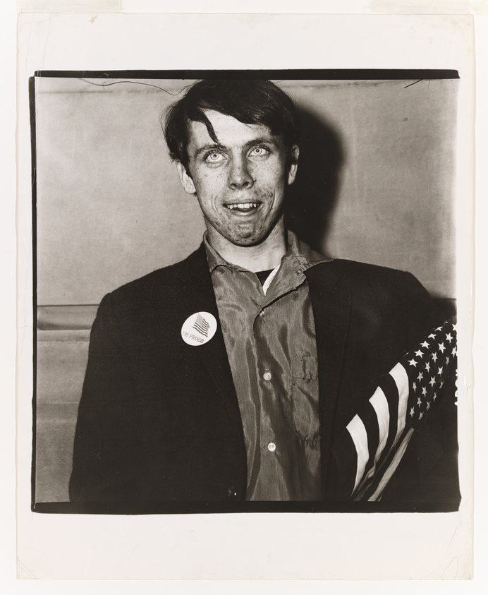 Black and white photo of a man with an exaggerated expression, wearing a button and holding an American flag.