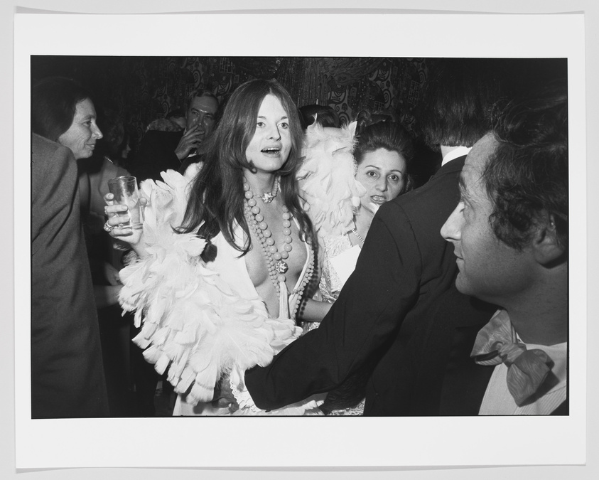 A black and white photograph capturing a lively social scene with a woman in the center wearing a feather boa and multiple necklaces, holding a drink in her hand, and smiling broadly. She is surrounded by other partygoers, including a man in the foreground looking towards her and a woman peeking out from behind him. The background features a patterned wallpaper and the atmosphere suggests a festive, crowded event.