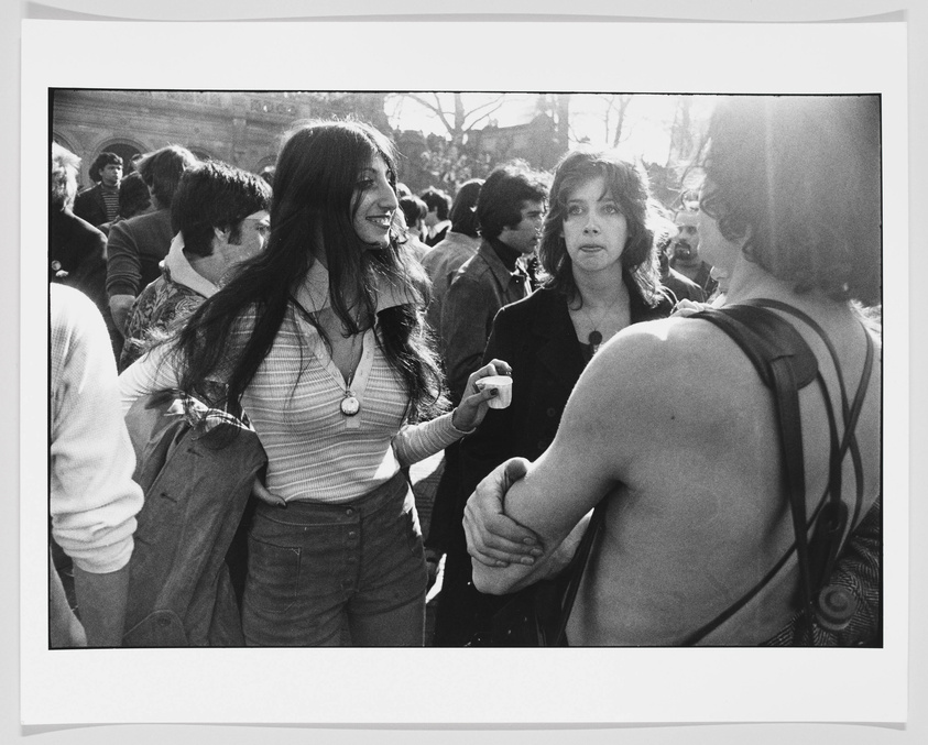 A black and white photograph capturing a candid moment among a group of people, likely from a past era. Two women are in the foreground, one smiling broadly with her hair flowing, the other looking on with a more reserved expression. They are surrounded by a crowd that suggests a social gathering or public event. The overall mood is lively and bustling.
