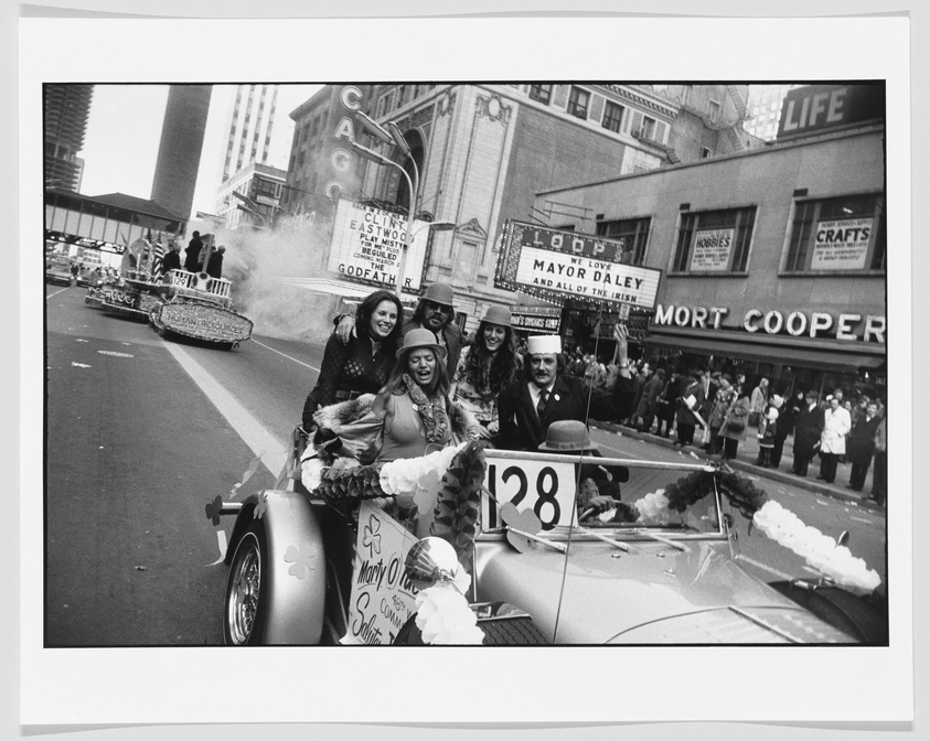 A black and white photo of a lively parade with people riding in decorated cars. The lead car, numbered 128, carries several individuals, including a woman in a festive outfit with a clover design, suggesting a St. Patrick's Day celebration. Spectators line the street, and signs for businesses like "LIFE" and "MORT COOPER" are visible in the background, along with a marquee supporting Mayor Daley. Smoke or steam can be seen rising from a float in the background.