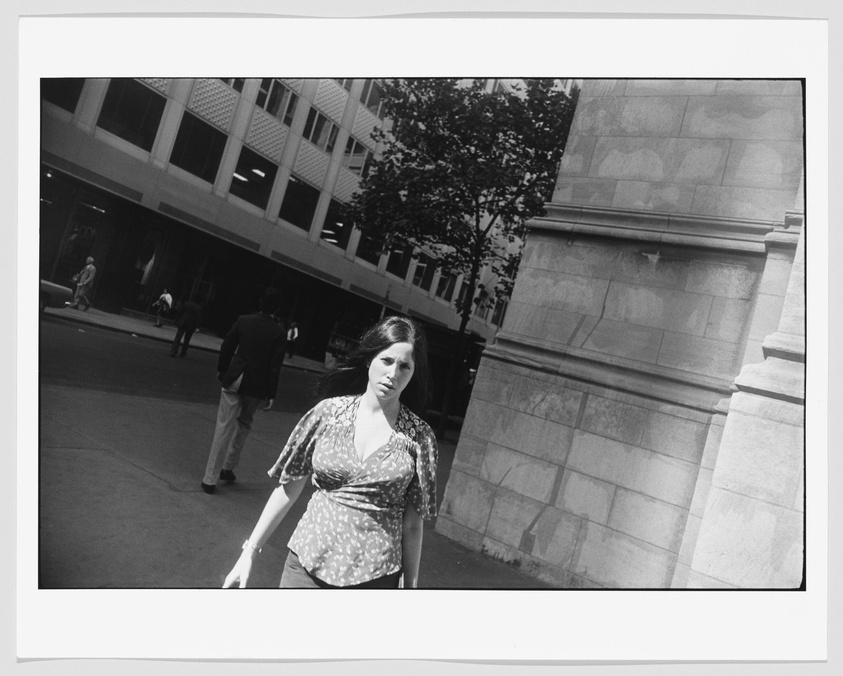 A black and white photograph capturing a woman walking towards the camera with a slightly concerned expression on her face. She is in the foreground on a city street with buildings, pedestrians, and trees in the background. The image has a candid, street photography feel to it.