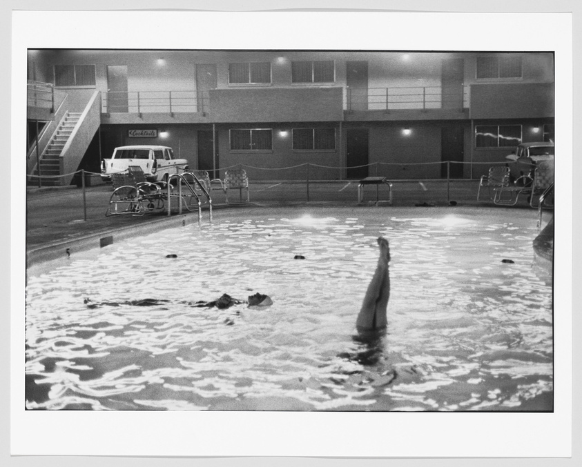 A black and white photograph depicting a nighttime scene at a motel swimming pool. Several people are swimming in the illuminated water, with one person's leg raised above the surface in the foreground. The two-story motel building in the background has its lights on, and a classic pickup truck is parked beside the pool area. There are lounge chairs and a staircase leading to the upper level of the motel. The atmosphere is quiet and still, with a sense of calm leisure.