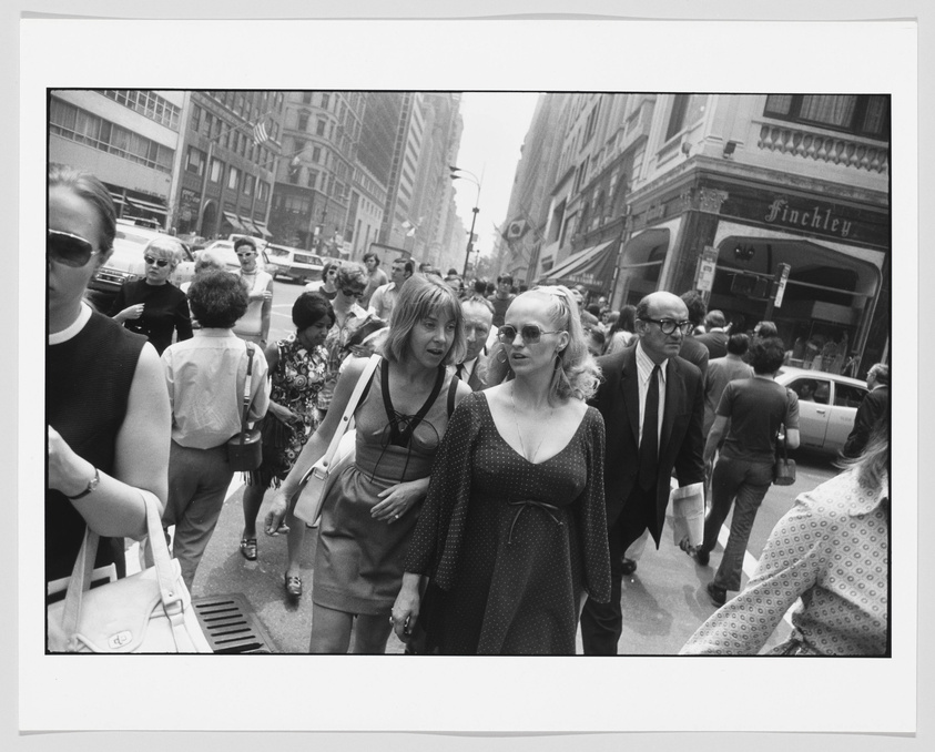 Black and white photograph of a bustling city street scene with pedestrians walking in various directions. Two women in the foreground appear to be conversing while walking. One woman is wearing sunglasses and a polka dot dress, and the other is in a sleeveless top and skirt with a shoulder bag. A man in a suit with glasses is also prominently featured in the center. The background shows a mix of 20th-century architecture, signage for a place called "Finchley," and cars parked along the street.