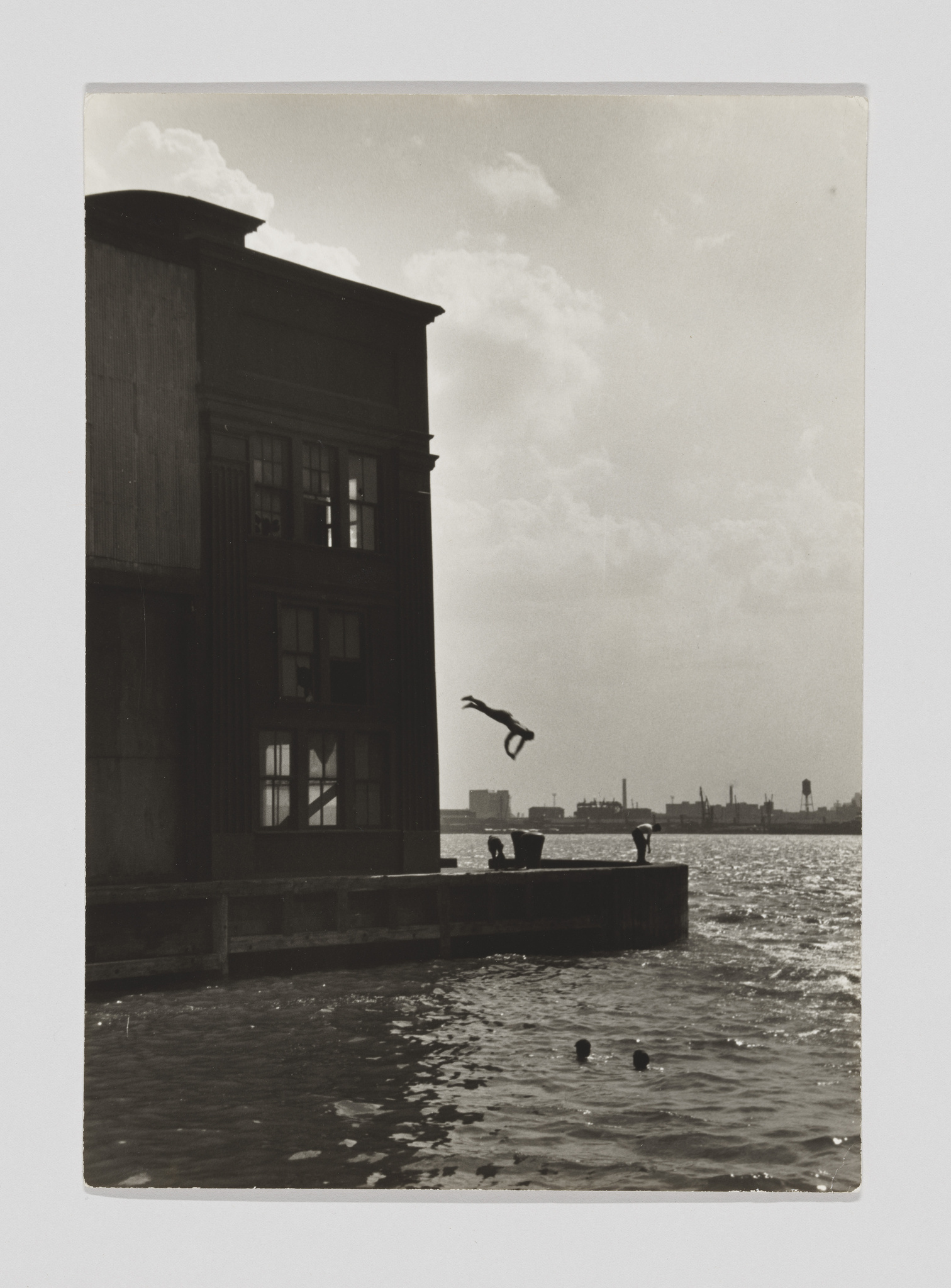 Person diving off a building into the water, with two others swimming nearby.