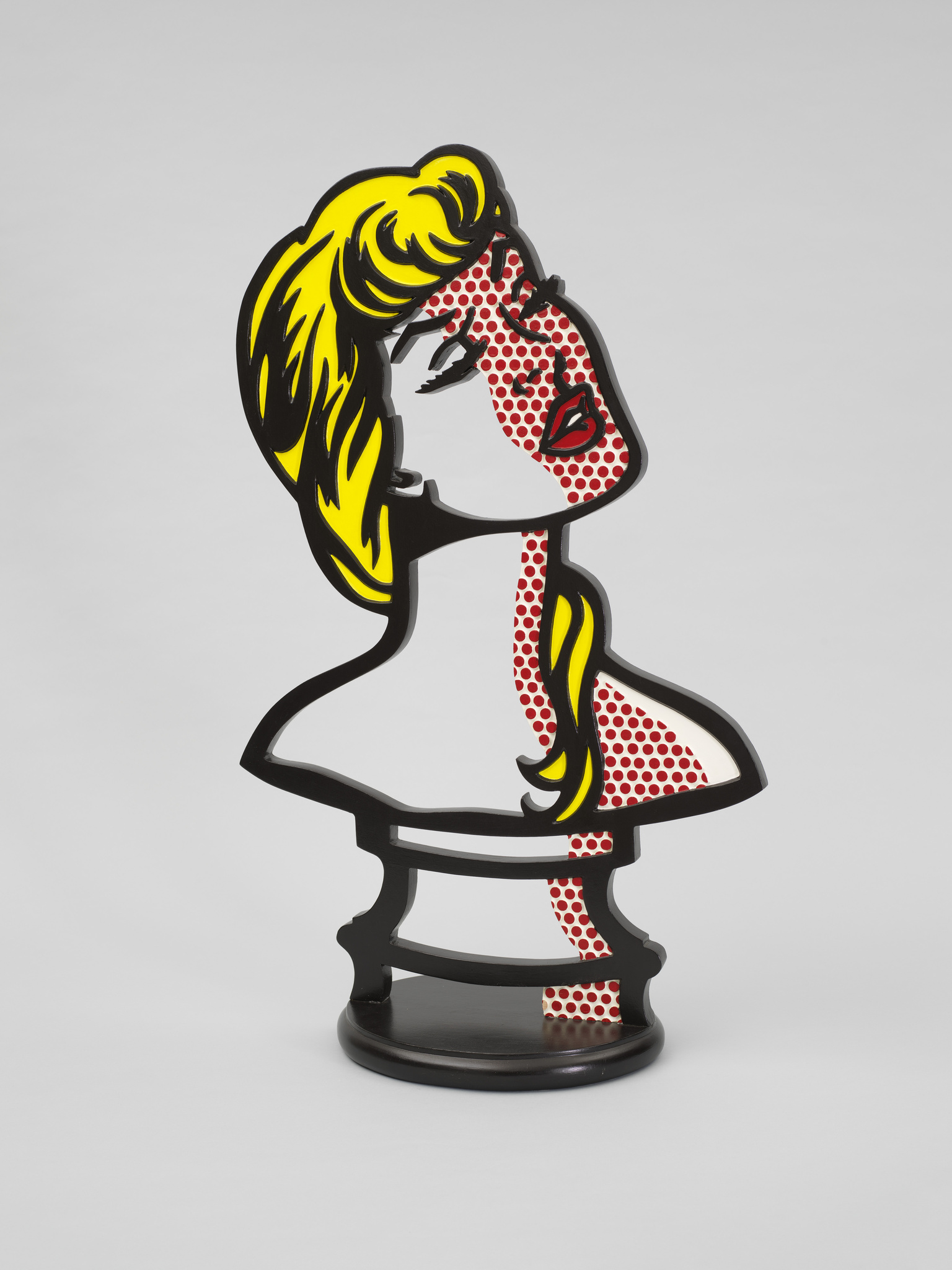 A stylized, three-dimensional representation of a female figure with pop art influences, featuring a bold black outline, yellow hair, and red dots on a white face and body, set against a plain background.