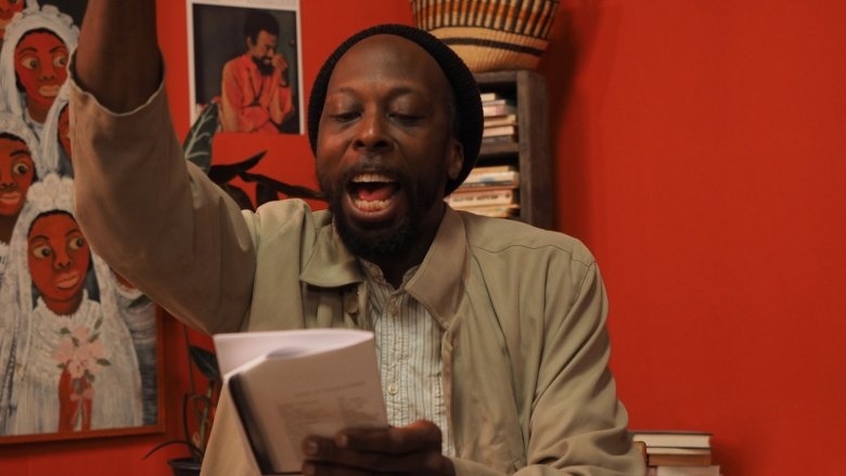 A man with a beanie hat is enthusiastically reading from a book, raising his left arm, with a vibrant red wall behind him adorned with colorful artwork.