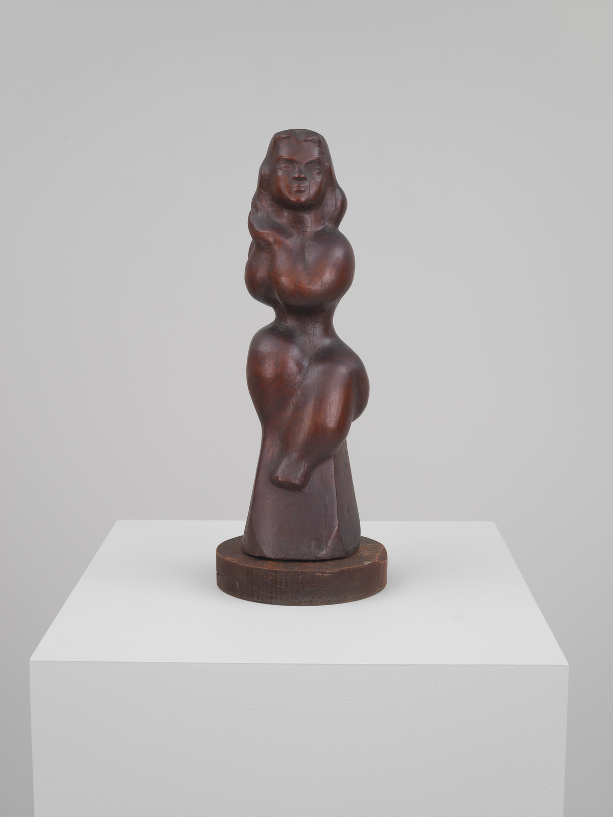 A wooden sculpture of a stylized female figure displayed on a white pedestal against a neutral background. The figure has smooth, flowing lines and simplified features, with an emphasis on the curves of the body.