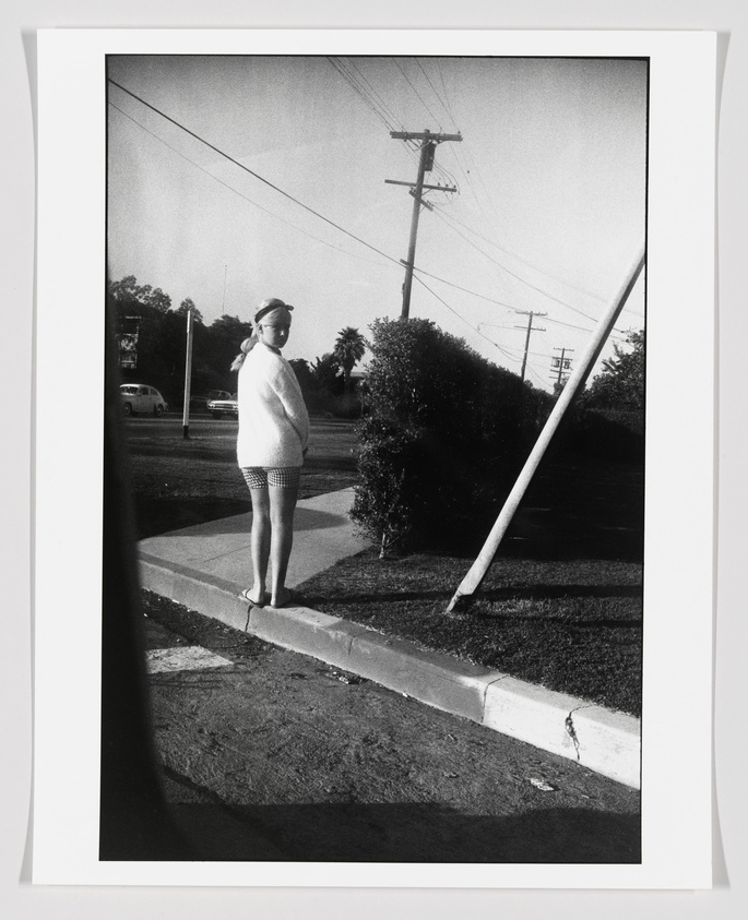 A black and white photograph depicting a woman standing on a sidewalk corner, wearing a light-colored sweater and patterned shorts, with her back partially turned to the camera. She is looking off to the side, with a residential street scene in the background, including a utility pole and power lines. The image has a vintage feel, possibly from the mid-20th century.