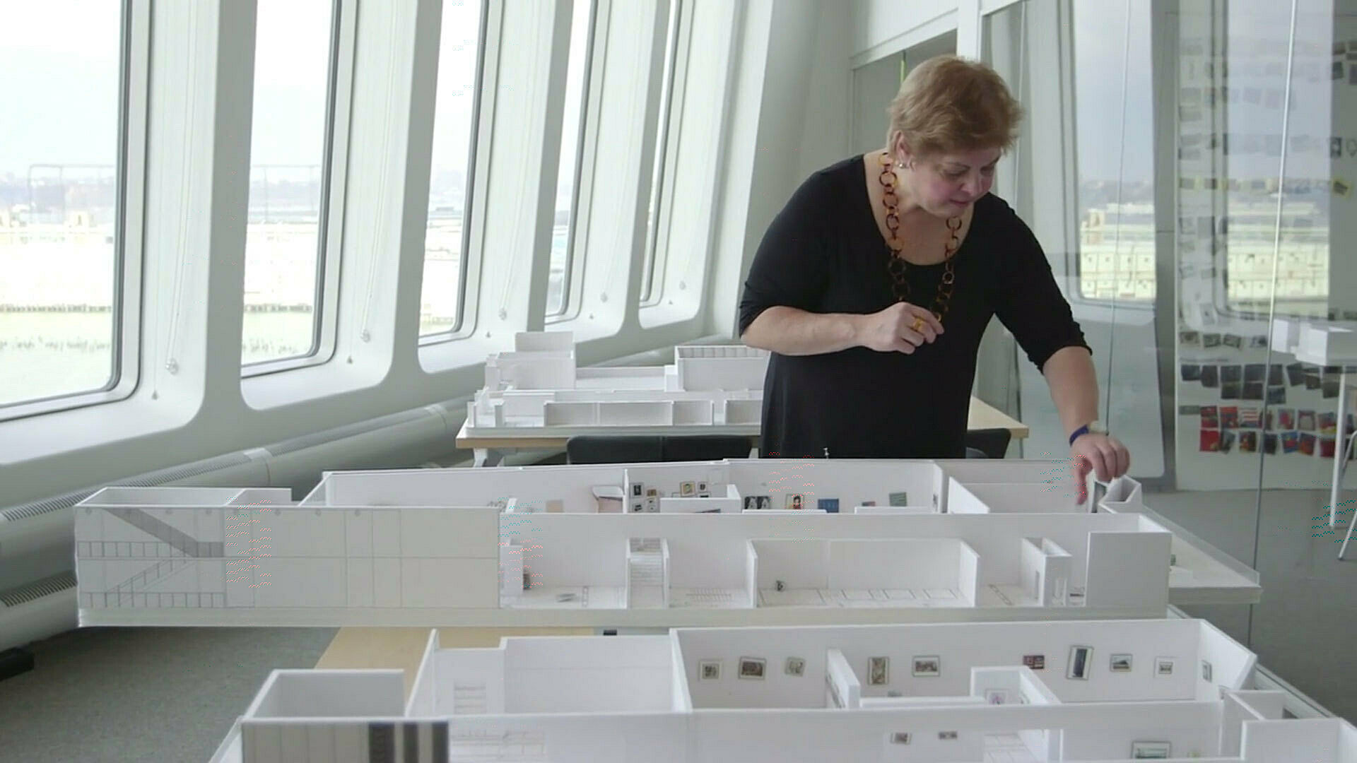A woman in a black top examines a detailed architectural model in a bright room with large windows and glass walls.