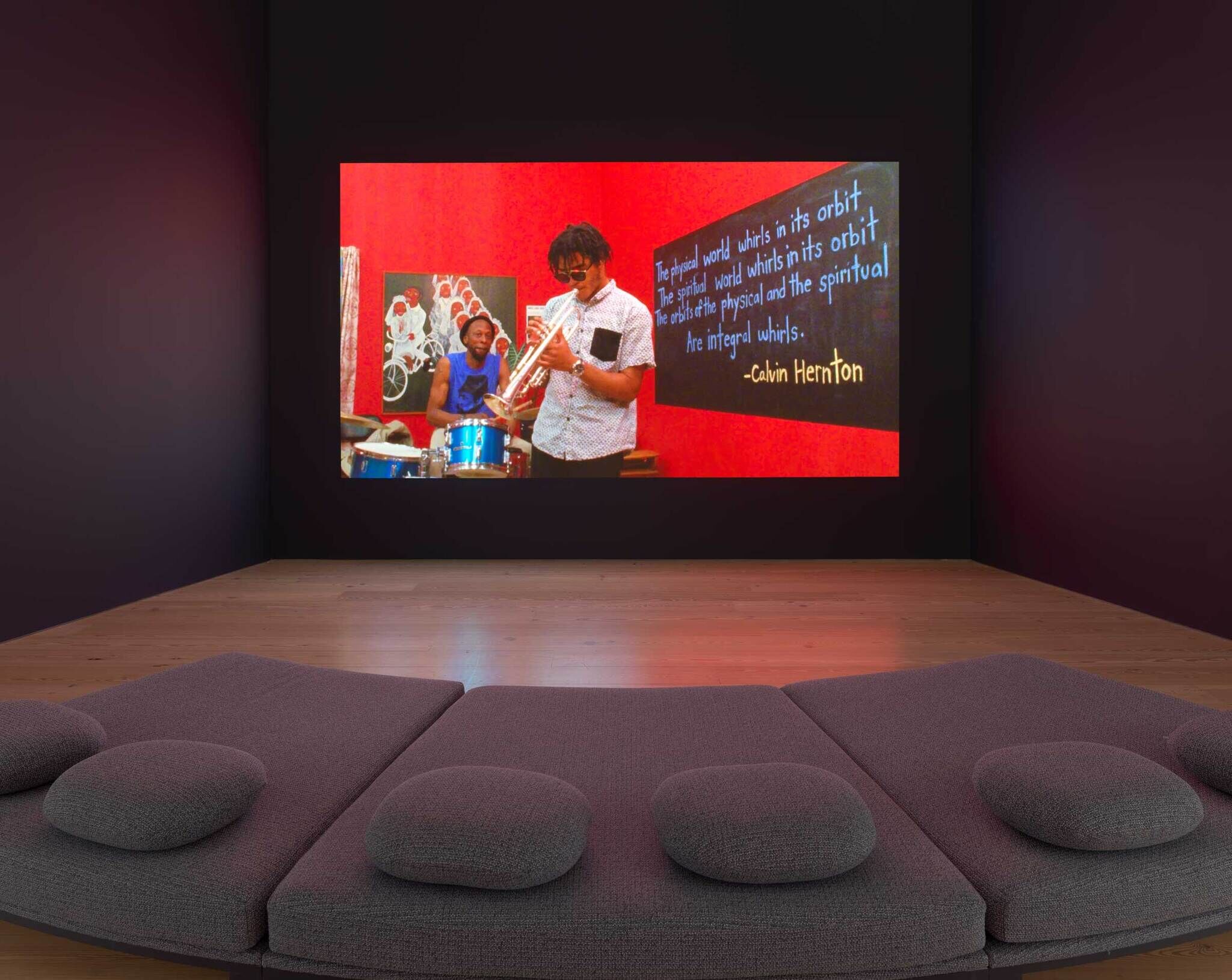 A grey couch and pillows pointing towards a big projection of a man playing the trumpet against a red background.